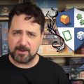 Ryan Henson Creighton makes board game videos for his YouTube channel, Nights Around a Table. Image depicts Ryan, a bearded man in his 40's, standing in front of a shelf full of various board games, next to the blue and white shield logo for Nights Around a Table